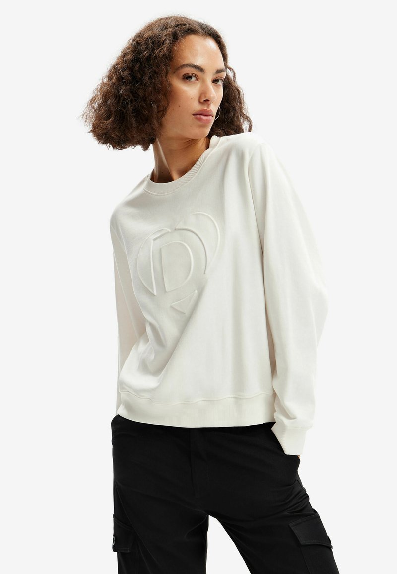 Sudadera blanca con un logo texturizado en el centro. Presenta un cuello redondo y mangas largas con puños. Combinada con pantalones cargo negros.