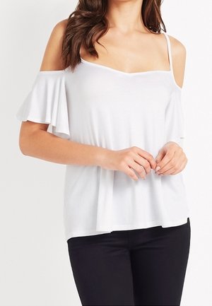 Mujer con una blusa blanca de hombros descubiertos con tirantes finos y pantalones negros, manos suavemente entrelazadas a la altura de la cintura.