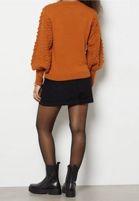 Pull couleur rouille avec des manches bouffantes texturées, associé à une mini-jupe noire et des bottines noires, sur un fond neutre.