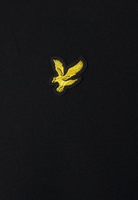 Camicia nera a maniche corte in tessuto testurizzato con un logo di aquila ricamato giallo sul lato superiore sinistro.
