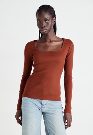 Kvinne med flette hår iført en rustfarget langermet topp med firkantet hals og lyseblå jeans, stående mot en ensfarget bakgrunn.