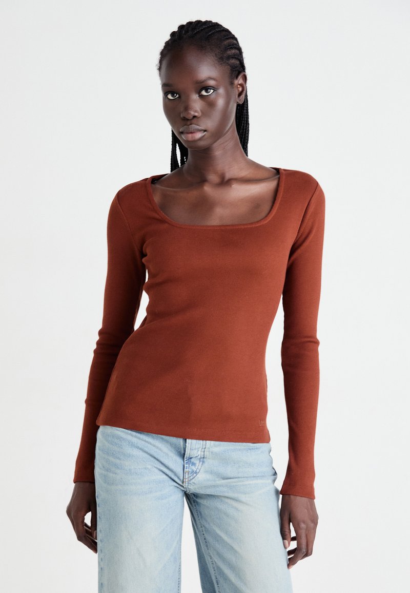 Vrouw met gevlochten haar die een roestkleurige lange mouw vierkante hals top en lichtblauwe jeans draagt, staand tegen een effen achtergrond.