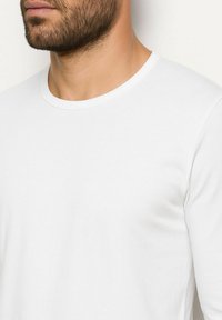 Weißes Langarm-T-Shirt mit Rundhalsausschnitt. Der Stoff wirkt glatt und leicht, sitzt eng an und hat keine auffälligen Muster oder Verzierungen.