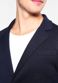 Navy blazer with a textured finish, notched collar, and a single chest pocket, paired with a white crewneck t-shirt underneath.