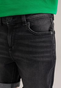 Črne jeans kratke hlače z zbledelim videzom, z gumbom za zapiranje, petimi žepi in zavihanimi manšetami, v kombinaciji z zeleno srajco.
