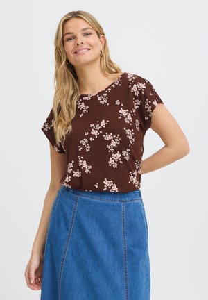Femme souriante aux longs cheveux blonds portant des boucles d'oreilles anneaux dorés, un haut marron fleuri à manches courtes et une jupe en jean bleue.