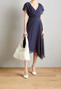 Marinblå omslagsklänning med korta fladderärmar, svepande fåll, i sällskap av en vit hobo-väska och vita spetstövletter. Slät tygtextur.