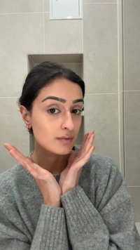 Gray knitted sweater, smooth skin, dark hair, hands framing the face, neutral background with light-colored tiles.