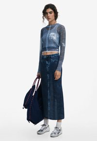 Blue knit long-sleeve top with graphic, high-waisted denim maxi skirt, paired with silver sneakers and a navy blue backpack with purple accents.