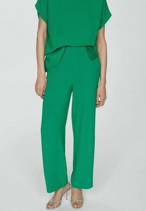 Vrouw draagt felgroene wijde broek en bijpassende top, gecombineerd met zilverkleurige sleehakken met bandjes, staand tegen een effen achtergrond.