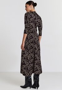Abito lungo e aderente in paisley marrone scuro con motivi dorati e bianchi, con maniche a tre quarti e orlo svasato. Stivali neri al ginocchio.