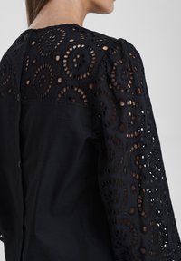 Donna che indossa una blusa nera con un ricamo a intaglio circolare intricata sulle maniche e sulla parte superiore della schiena, dotata di chiusura con bottoni lungo la parte centrale della schiena.