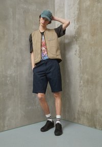 Gilet sans manches beige sur un t-shirt graphique, shorts navy, chaussures noires et chaussettes rayées. Fond texturé dans des nuances de gris.