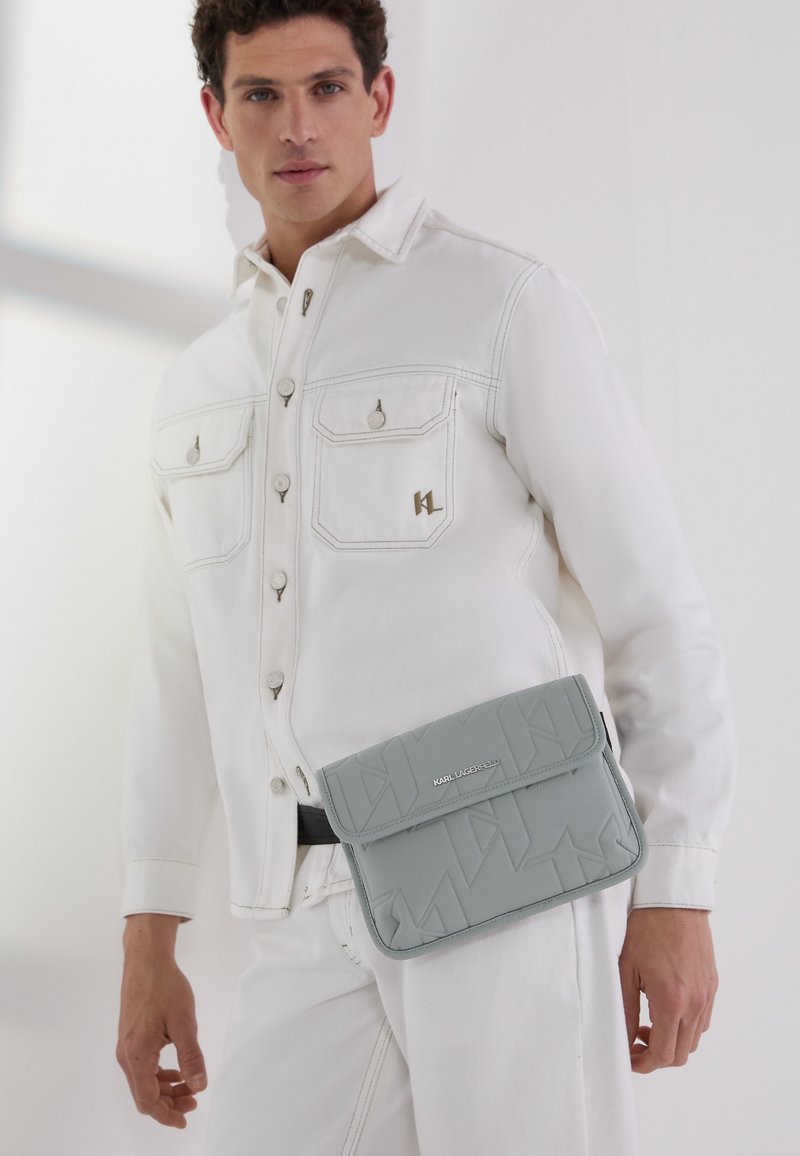 Borsa a tracolla in pelle grigia con texture, dotata di chiusura a pattina, caratterizzata da un design geometrico e un dettaglio del logo metallico. Indossata con una giacca bianca.