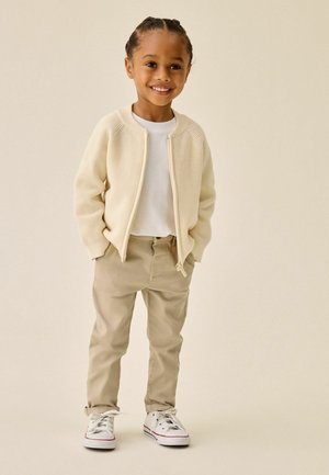 Young child with braided hair smiling, wearing a cream knit zip-up sweater, white shirt, beige pants, and white sneakers standing with hands in pockets.