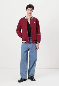 Bordo varsity jakna s crno-bijelim prugastim rebrastim ovratnikom i manžetama, s pritisnim dugmadima, nosi se preko crne majice i svijetloplavih traperica.