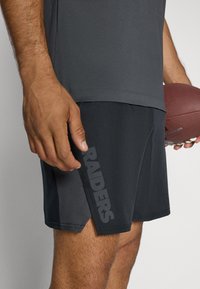 Calças curtas atléticas pretas com uma abertura lateral, apresentando um grande logótipo cinza "RAIDERS". A textura é suave, feita com um material leve que absorve a humidade.