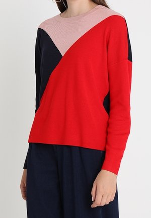 Femme portant un pull à blocs de couleurs avec des sections rouges, noires et roses, associé à un pantalon ample bleu marine foncé, debout devant un fond blanc.