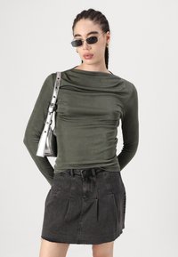 Oliven grønn langermet topp med drapert halslinje, parret med et svart denim miniskjørt med legg og uferdige kanter. Sølvfarget håndveske og solbriller.