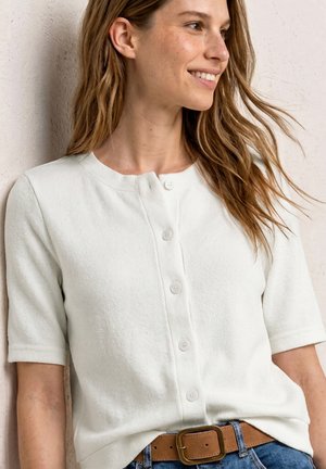 Femme souriante aux longs cheveux bruns portant un cardigan blanc à manches courtes boutonné et un jean bleu avec une ceinture marron.
