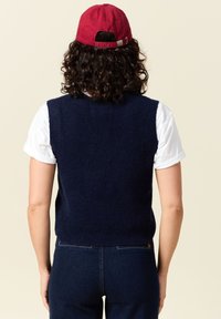 Marinblå, ärmlös stickad väst i mjukt material, parat med en vit kortärmad skjorta, bärs med mörk denimjeans och en röd keps.