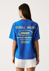 Zila kokvilnas T-kreklā ir uzdrukāts "RED BULL RACING" un "DRIVEN BY PERFORMANCE" grafikas dzeltenā krāsā uz muguras. Īsās piedurknes un brīva piegriezuma.