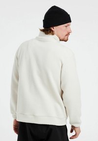 Hvid fleece-pullover med høj krave og lange ærmer. Blød tekstur, afslappet pasform og ribbet kant. Sort hue bæres på hovedet.