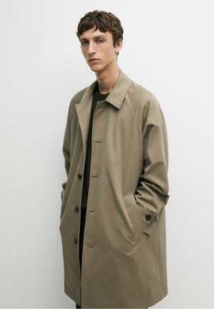Giovane uomo con capelli corti e ricci indossa un cappotto color cammello con bottoni e mani nelle tasche, in piedi davanti a uno sfondo chiaro e semplice.