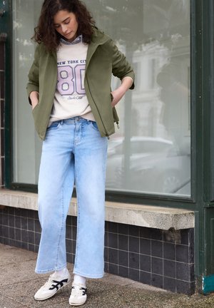 Nainen, jolla on vaaleansiniset leveälahkeiset farkut, valkoiset tennarit, luonnonvalkoinen collegepaita, jossa lukee "New York City 80", ja oliivinvihreä takki, seisomassa ulkona.