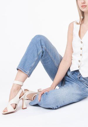 Sandali con tacco alto beige, con design incrociato, cinturino alla caviglia e tacco in legno, abbinati a jeans in denim di un blu chiaro e una camicia bianca con bottoni.