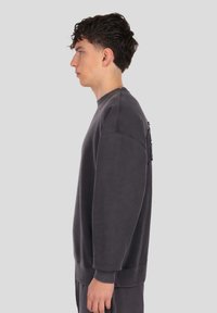 Szara bluza z zaokrąglonym dekoltem, długimi rękawami, ściągaczami na mankietach i dolnym brzegu; posiada teksturowaną naszywkę po lewej stronie. Miękki, casualowy materiał.