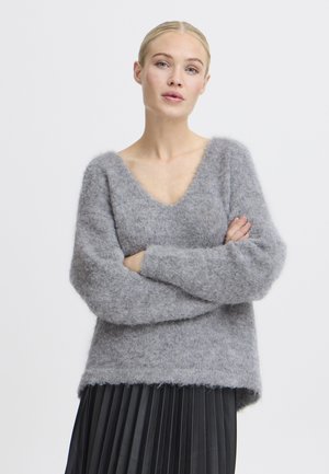 Pull gris flou avec un large col en V, épaules tombantes et tissu texturé. Associé à une jupe plissée noire.