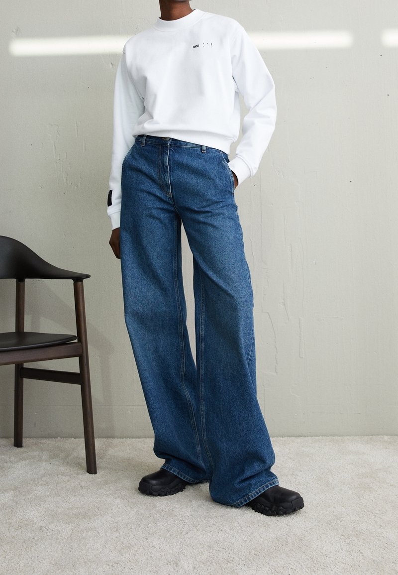 Personne portant une chemise blanche à manches longues, un jean bleu ample taille haute et des chaussures noires, debout à côté d'une chaise en bois sur un tapis.