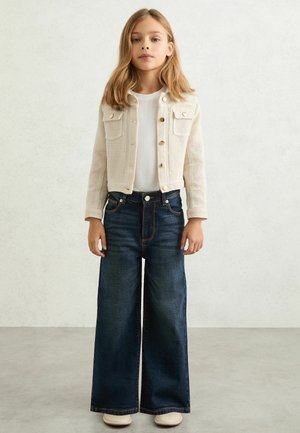 Beżowy prążkowany kardigan z kieszeniami, biała koszulka, szerokie ciemne dżinsy oraz jasne buty, stojąca na tle o fakturze.