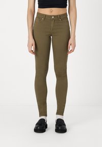 Pantalones vaqueros ajustados de color verde oliva con cintura de altura media, diseño de cinco bolsillos y detalles de costuras en contraste. Combinados con zapatos negros y calcetines blancos.