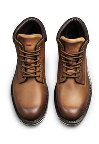 Bottes en cuir marron avec bouts ronds, fermeture à lacets, col rembourré et détails de surpiqûres contrastantes. La semelle présente un motif de crampons robuste.