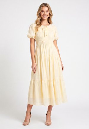 Lachende vrouw in een lichtgele, korte mouw, gelaagde midi-jurk met een striksluiting aan de halslijn en beige sandalen met hoge hakken.