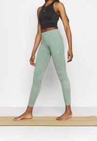 Top sportivo nero con dettagli in rete abbinato a leggings high-waisted verde chiaro con particolari testurizzati. Il modello è in piedi a piedi nudi su un tappetino.