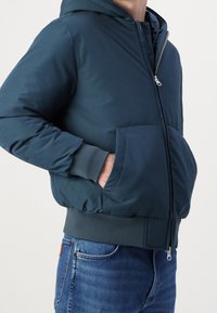 Tmavě modrá bunda s kapucí, elastickými manžetami a pasem, s předním zipem a velkými bočními kapsami. Hladká textura tkaniny.
