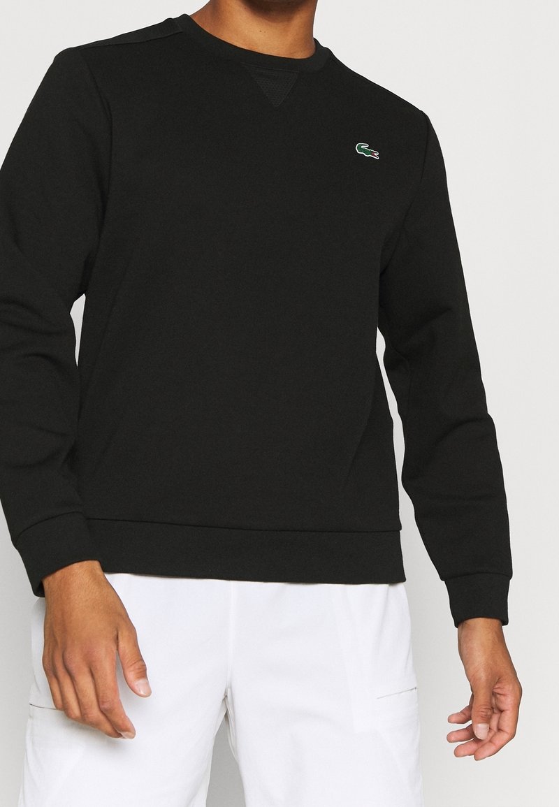 Homme portant un sweat-shirt noir à manches longues avec un petit logo de crocodile vert sur la poitrine et un pantalon blanc.