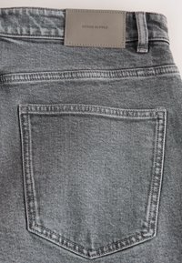 Aizmugurējā kabata pelēkajām džinsa biksēm ar redzamu šuvumu un pelēkas ādas zīmola uzlāpi ar uzrakstu "DENIM SUPPLY" virs kabatas.