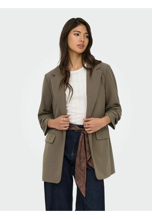Donna che indossa un blazer color taupe sopra una maglia bianca strutturata, jeans scuri a gamba larga e una sciarpa marrone fantasia legata in vita.