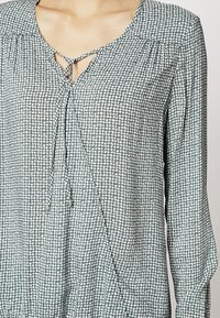 Długa koszula z długim rękawem w zielono-białym wzorze, z wiązaniem przy dekolcie w kształcie dziurki oraz marszczeniami na ramionach, które nadają kształt.