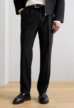 Άνδρας που φορά μαύρο ραμμένο παντελόνι, μαύρα δερμάτινα μοκασίνια, μαύρο σακάκι και λευκό πουλόβερ, στέκεται σε ξύλινο πάτωμα.