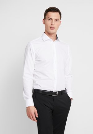 Weißes Langarmhemd mit einem Kentkragen; verfügt über ein gemustertes Innenkragen-Detail. Getragen mit schwarzen Hosen und einem schwarzen Gürtel.