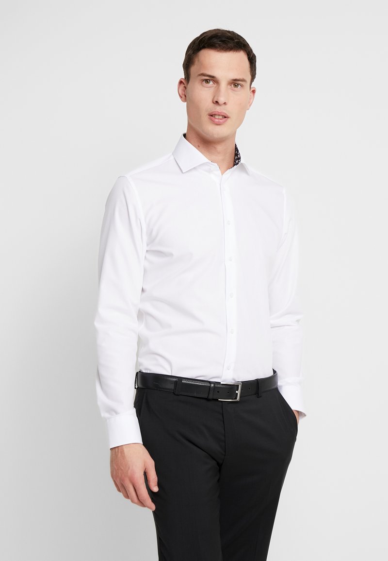 Camicia bianca a maniche lunghe con colletto cutaway; presenta un dettaglio interno del colletto a fantasia. Indossata con pantaloni neri e una cintura nera.