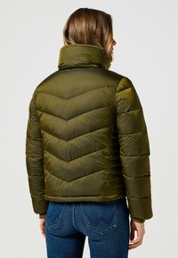 Oliwkowa kurtka puchowa z wysokim kołnierzem, w wzór chevron, o miękkiej fakturze i elastycznych mankietach. Noszona z niebieskimi dżinsami, prezentując casualowy fason.