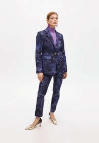Completo giacca e pantaloni di colore blu scuro con una stampa animale testurizzata, accentuato da un bottone dorato. Abbinato a un dolcevita viola chiaro e décolleté dorati.