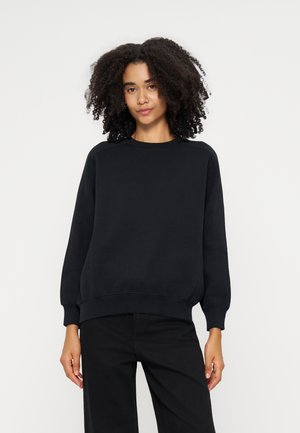 Femme aux cheveux bouclés portant un sweat-shirt noir uni à manches longues et un pantalon noir, debout devant un fond blanc.