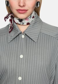 Graues gestreiftes Hemd mit großem Kragen und weißen Knöpfen. Ein bedruckter Schal mit Kirschmotiven ist um den Hals gebunden. Glatte Stoffstruktur.